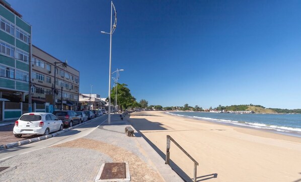 A coastal road with a beach, parked cars, and buildings. - Apto com Garagem a 550 Metros da Praia do Centro (Rio das Ostras)