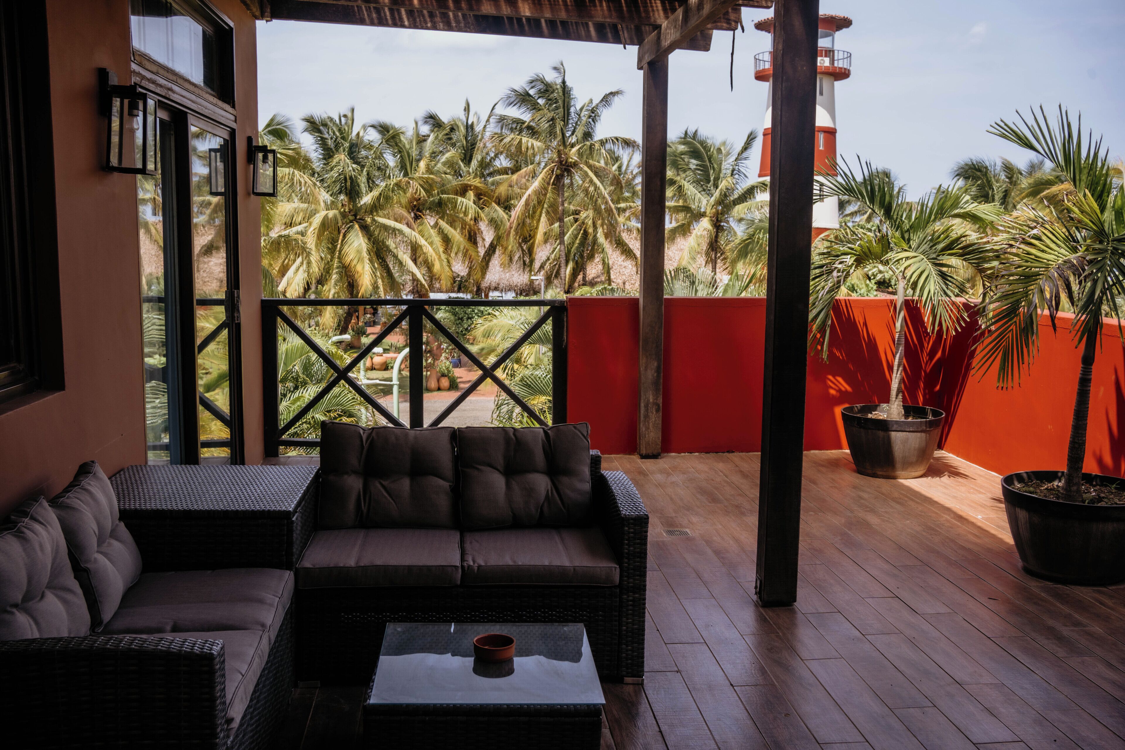 A covered outdoor seating area with a sofa and chairs, a table, and a view of palm trees and a red wall.