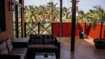 A covered outdoor seating area with a sofa and chairs, a table, and a view of palm trees and a red wall.