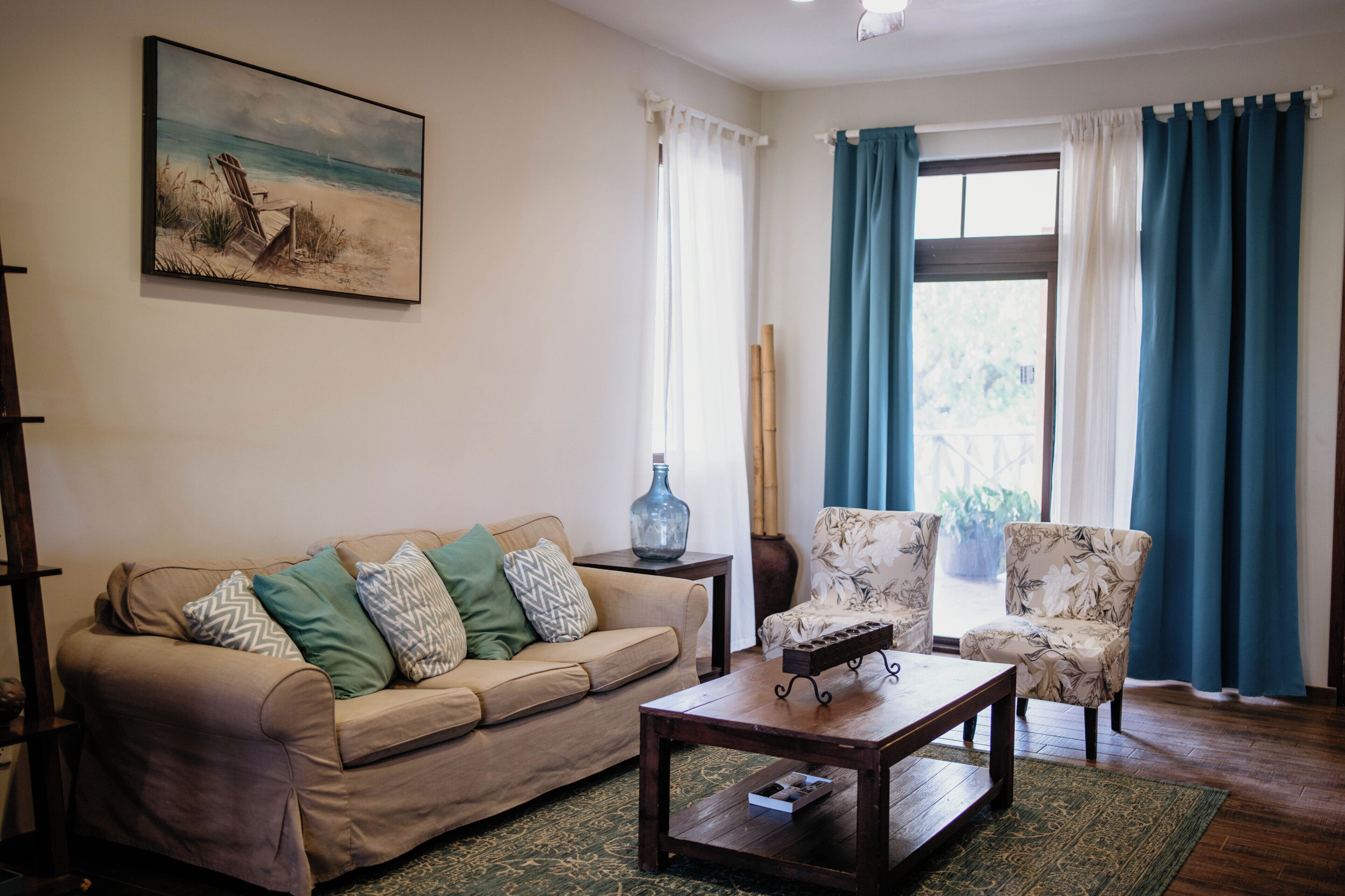 Ein Wohnzimmer mit einer Couch, einem Couchtisch und zwei Sesseln. An der Wand hängt ein Bild, ein Fenster mit Vorhängen und ein Holzboden.