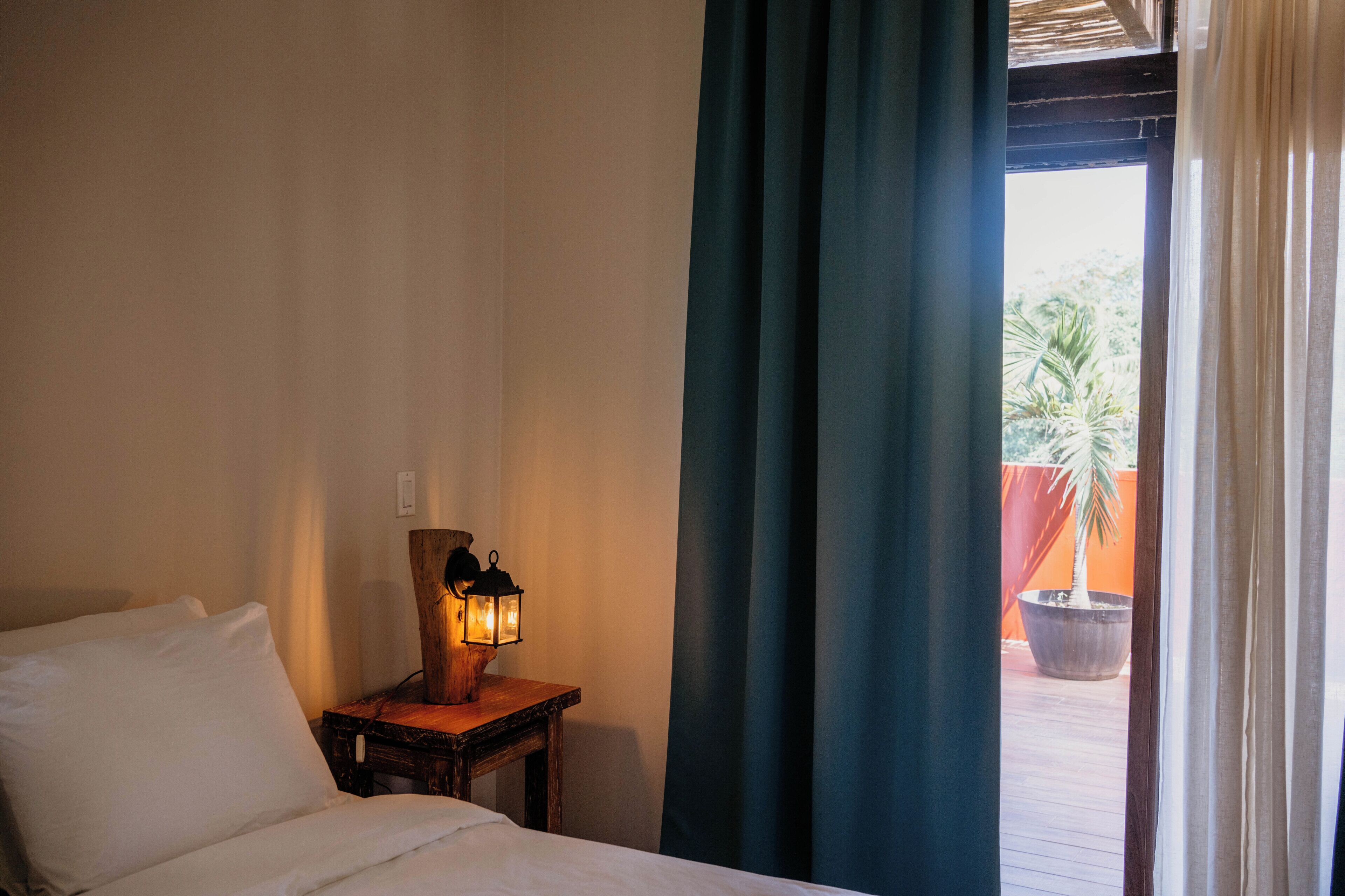 A hotel room with a bed, a nightstand with a lantern, and a view of a courtyard with a palm tree.