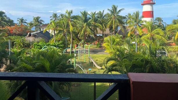 A view of a park with a playground, palm trees, and a lighthouse in the background.