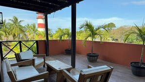 A covered outdoor seating area with wooden furniture and potted plants, overlooking a red wall and palm trees.