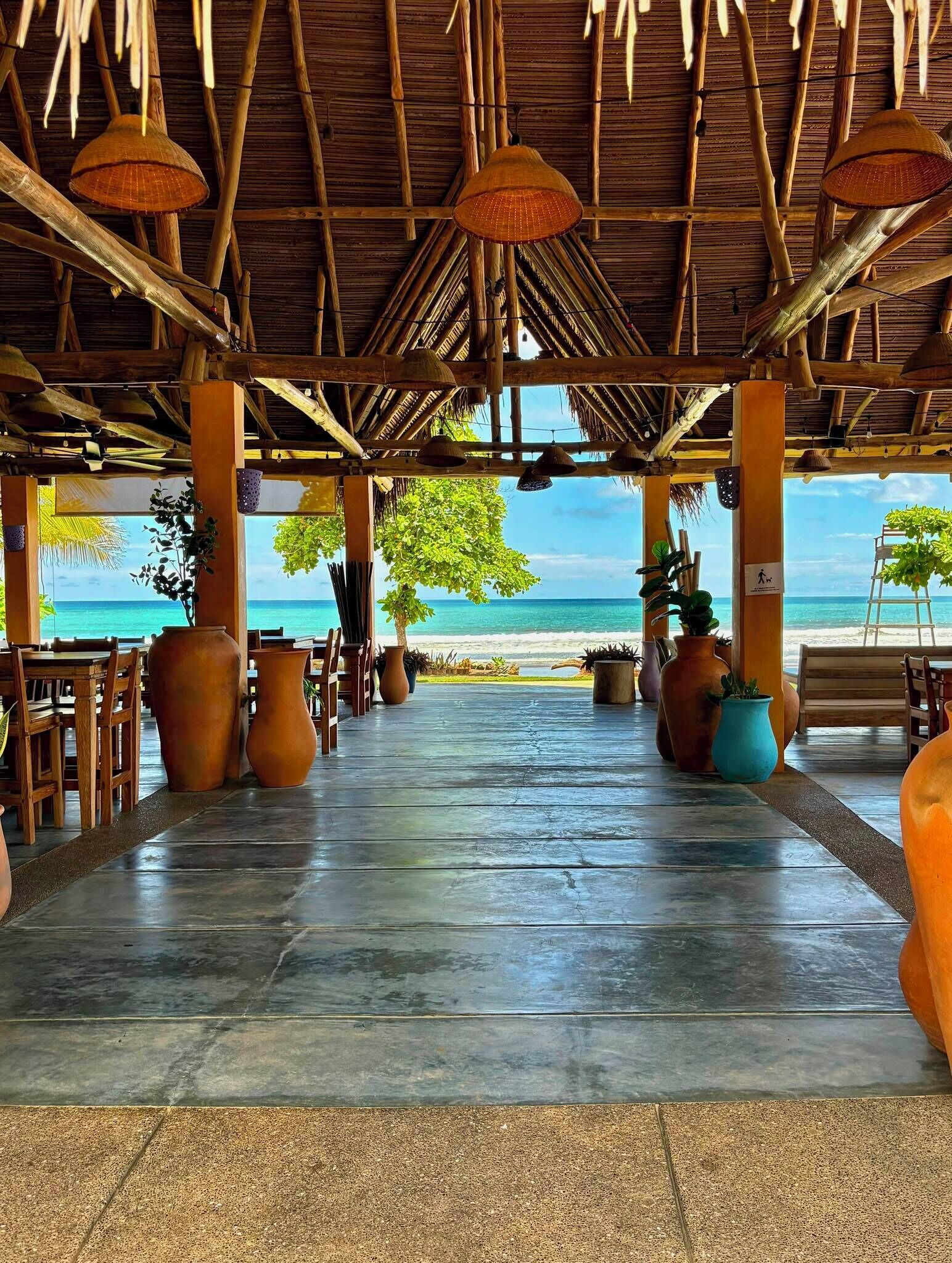An open-air dining area with a view of the beach, featuring a thatched roof, wooden furniture, and potted plants.