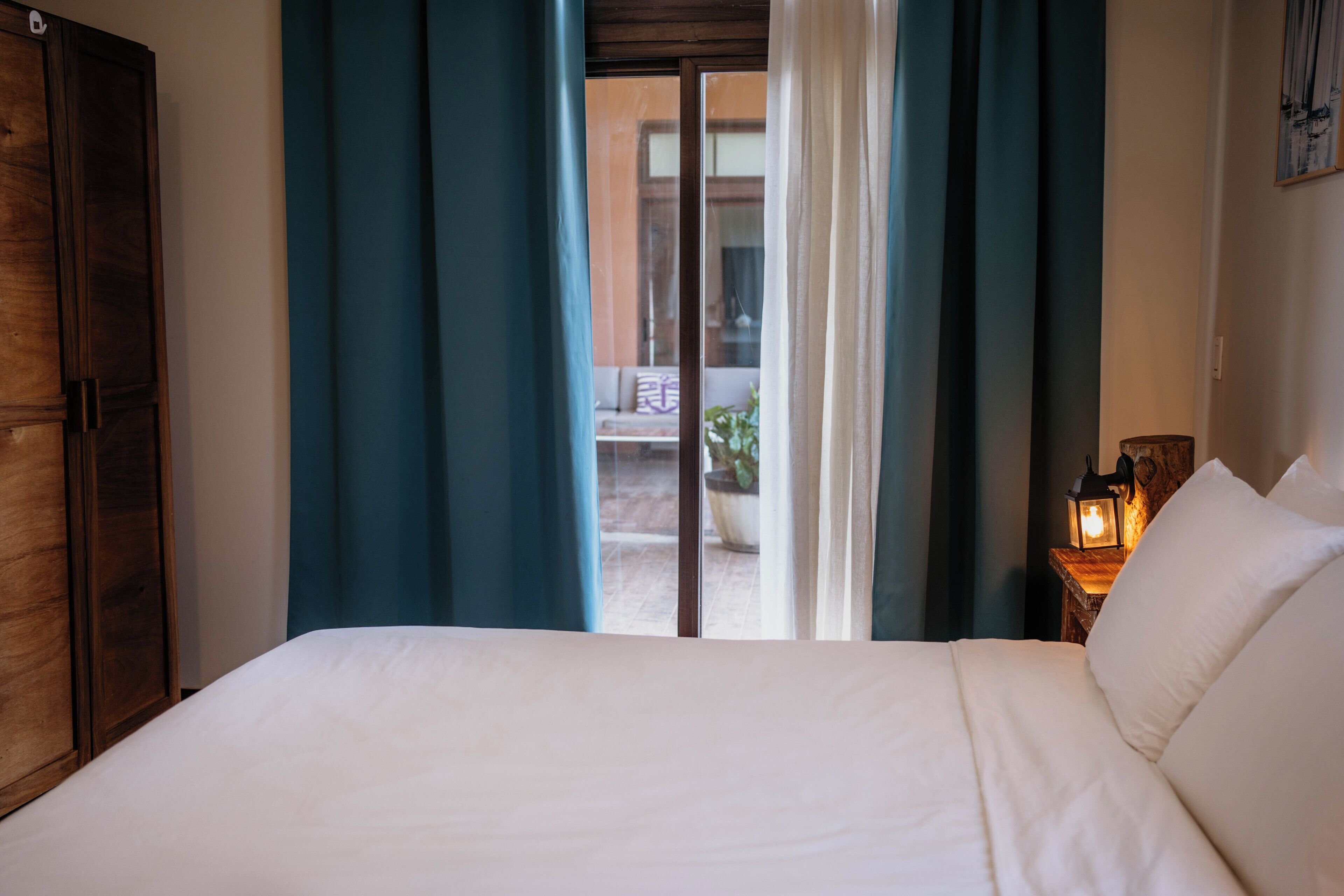 A hotel room with a bed, curtains, a wooden wardrobe, and a view through a glass door.