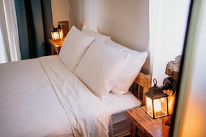 Una cama bien hecha con sábanas y almohadas blancas, un cabecero de madera y dos lámparas de noche sobre mesitas de madera.