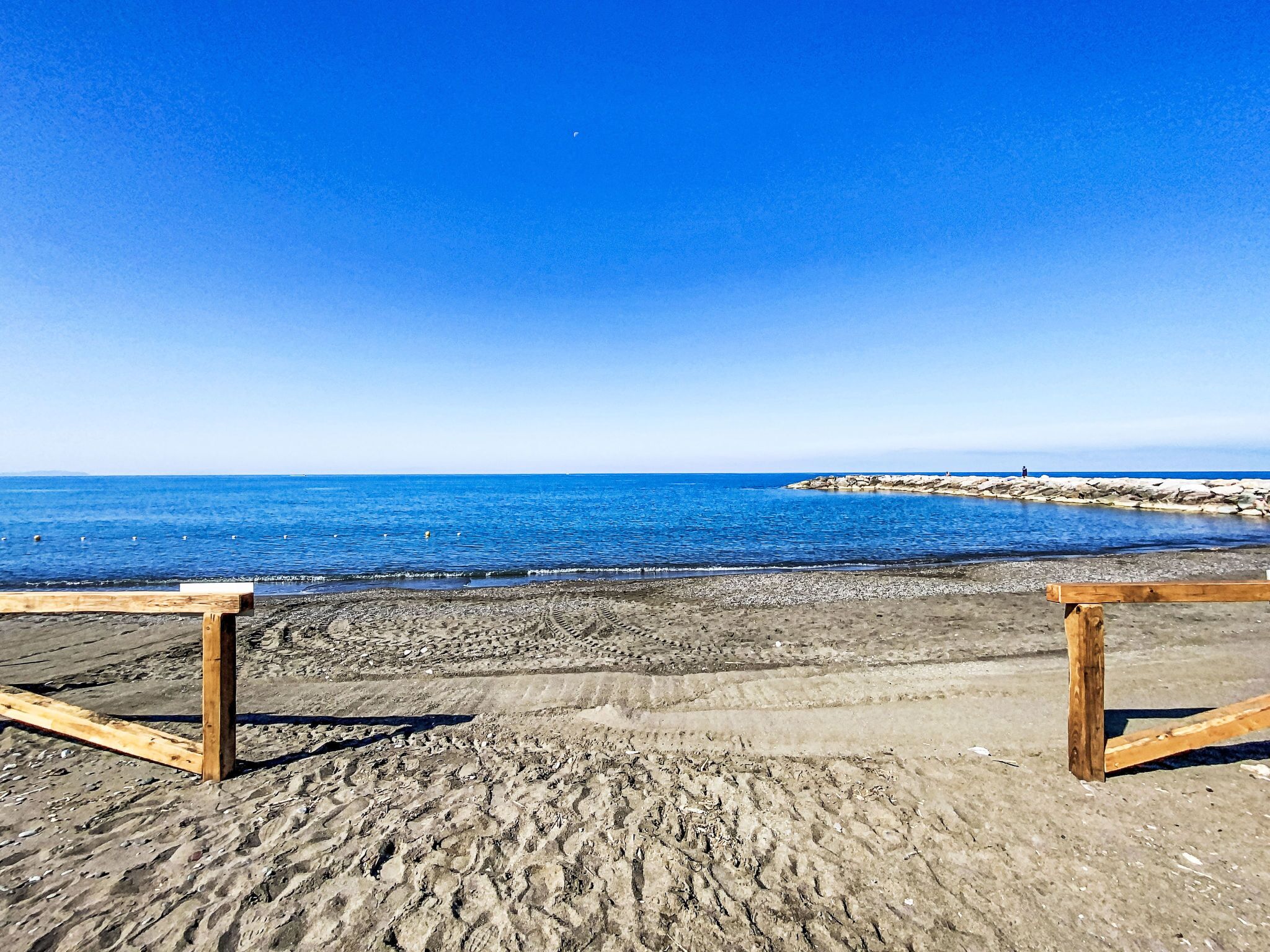 A beach with wooden benches, a rocky shoreline, and calm blue waters.