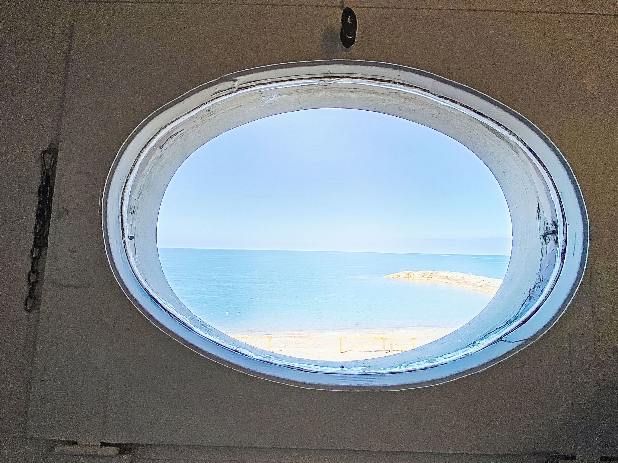 A round window with a view of the sea and a sandy beach.
