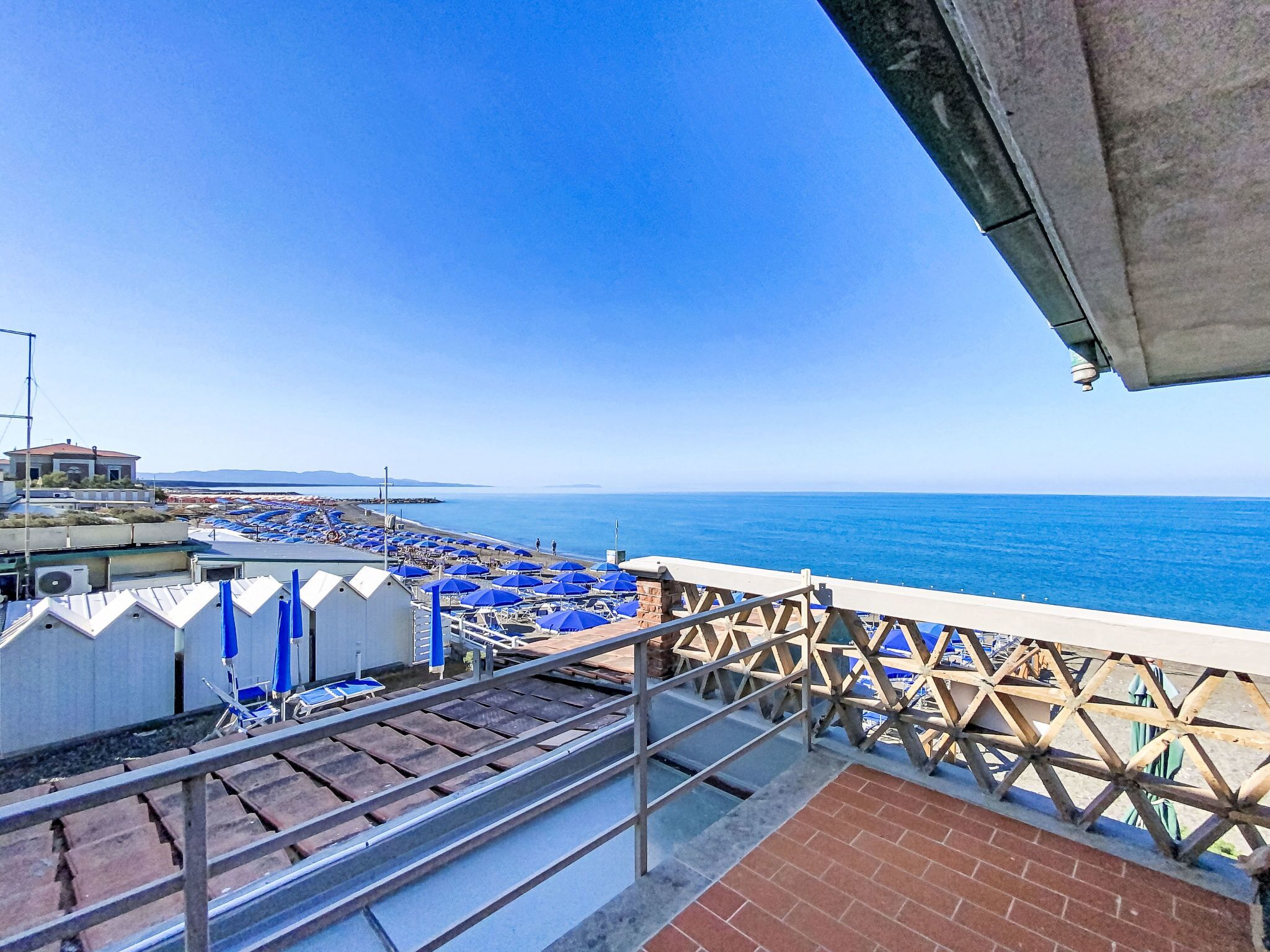 A balcony with a view of the sea, blue umbrellas, and a brick floor.