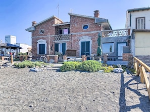 A two-story brick building with a balcony, a small garden area with rocks and plants, and a wooden fence. - Ginepro sul mare by Interhome (Cecina)