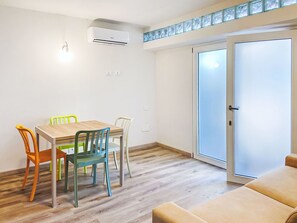 Een kamer met een houten eettafel, kleurrijke stoelen, een beige bank, een schuifdeur met glas en een airconditioner.