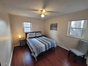 Ein Schlafzimmer mit einem Bett, einem Nachttisch, einem Sessel, einem Deckenventilator und zwei Fenstern.