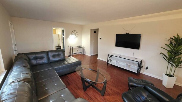 A living room with a sectional sofa, a glass coffee table, a flat-screen TV mounted on the wall, and a potted plant. - Remodeled Rossford Lower Apartment (Rossford)