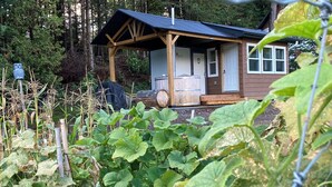 Une cabane en bois dotée d’un porche, entourée d’un jardin où poussent différentes espèces de plantes.