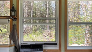 Une pièce avec un poêle à bois, un réfrigérateur, une fenêtre donnant sur les arbres et une étagère avec une lanterne.
