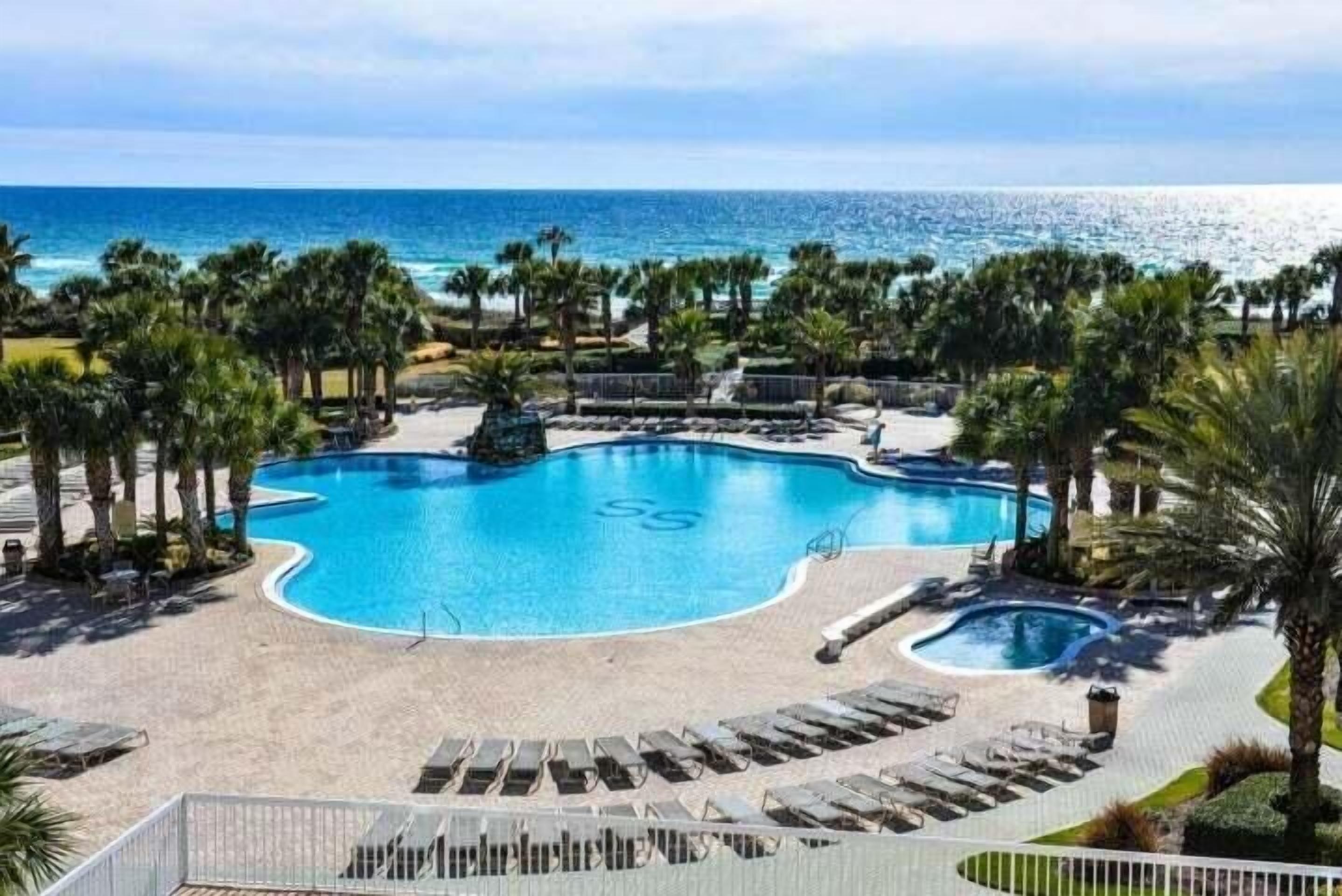 A resort pool with a unique shape, surrounded by palm trees and lounge chairs, with a view of the ocean.