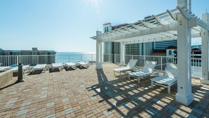 Eine sonnendurchflutete Terrassenanlage im Freien mit weißen Liegestühlen und einer Pergola, mit Blick auf das Meer.
