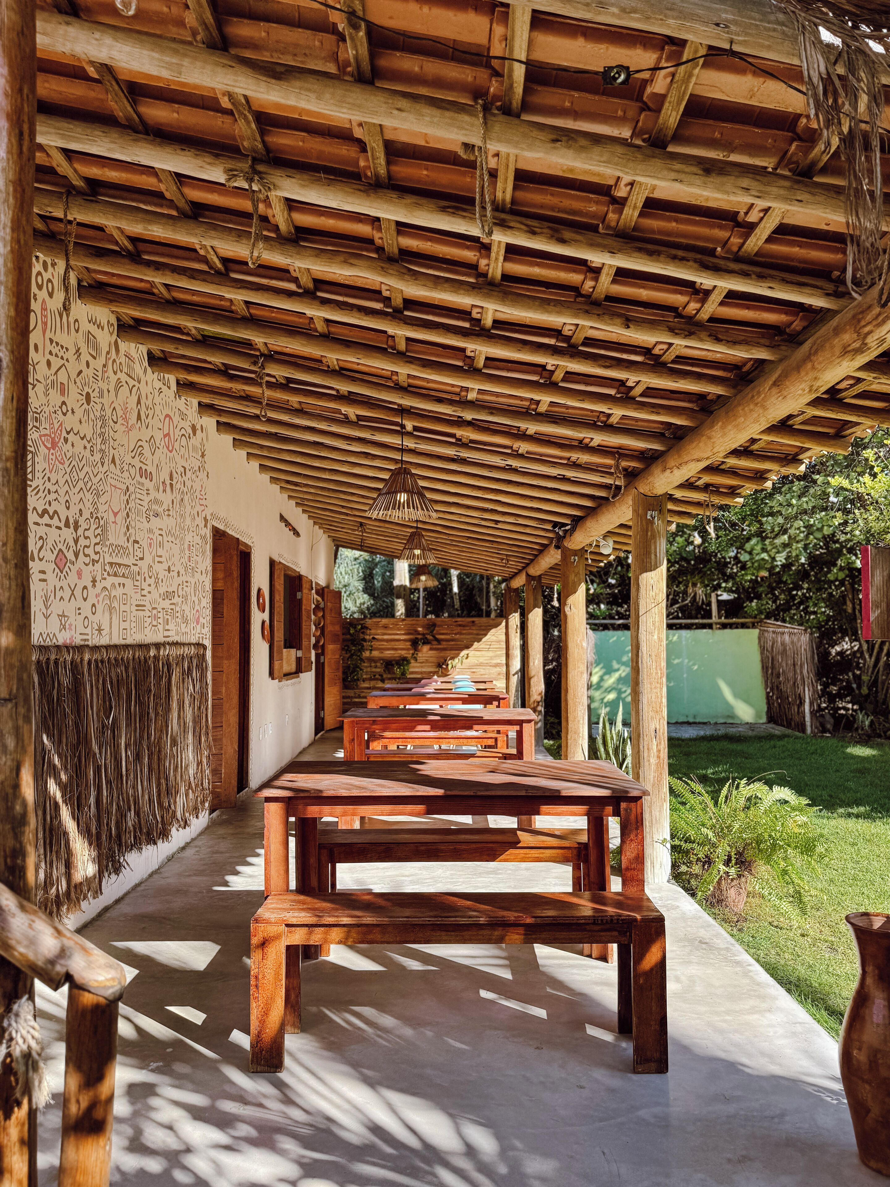 Un espacio al aire libre cubierto con bancos y mesas de madera, suelo de concreto y una pared con diseños decorativos.