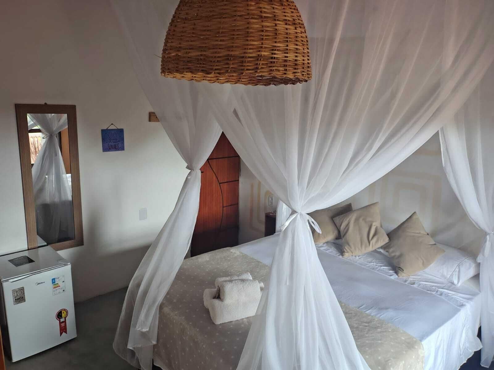 Un dormitorio con cama, cortinas blancas, dosel de mimbre, espejo y un pequeño electrodoméstico blanco.