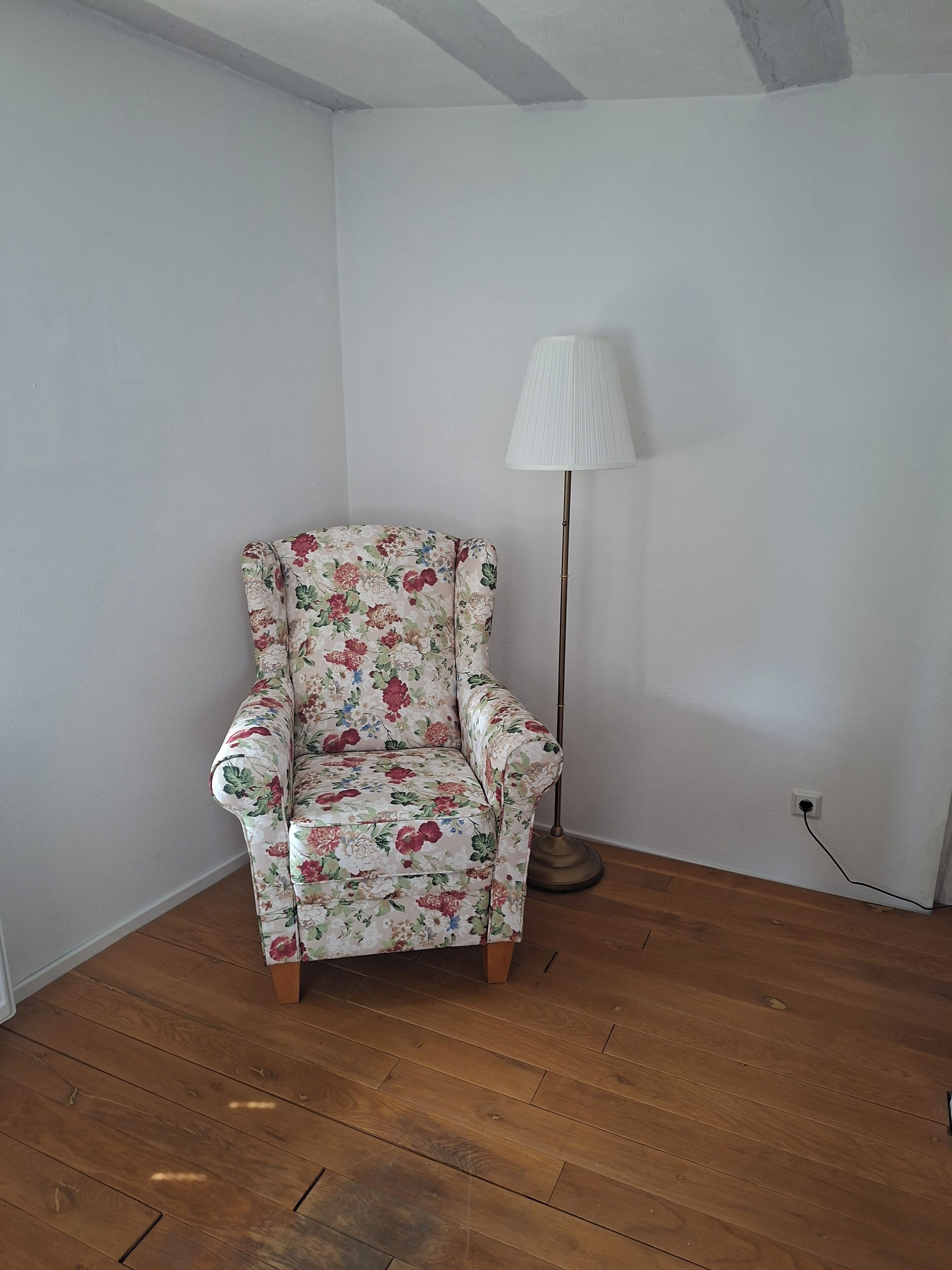 Une pièce d’angle avec un fauteuil à motifs floraux, un lampadaire et un plancher en bois.
