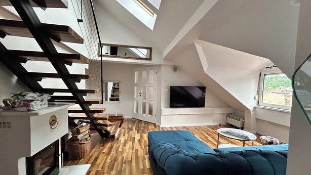 A loft-style room with a wooden floor, a blue sofa, a TV mounted on the wall, a wooden staircase, and a window with a view. - Spacious attic duplex in a heritage building in central Sarajevo (Sarajevo)