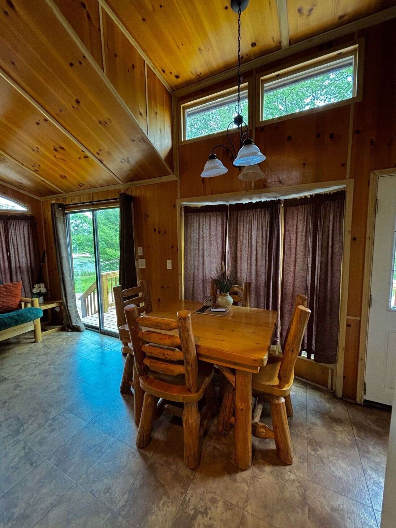 A wooden dining room with a large table and chairs, a hanging light fixture, and a view through the windows.