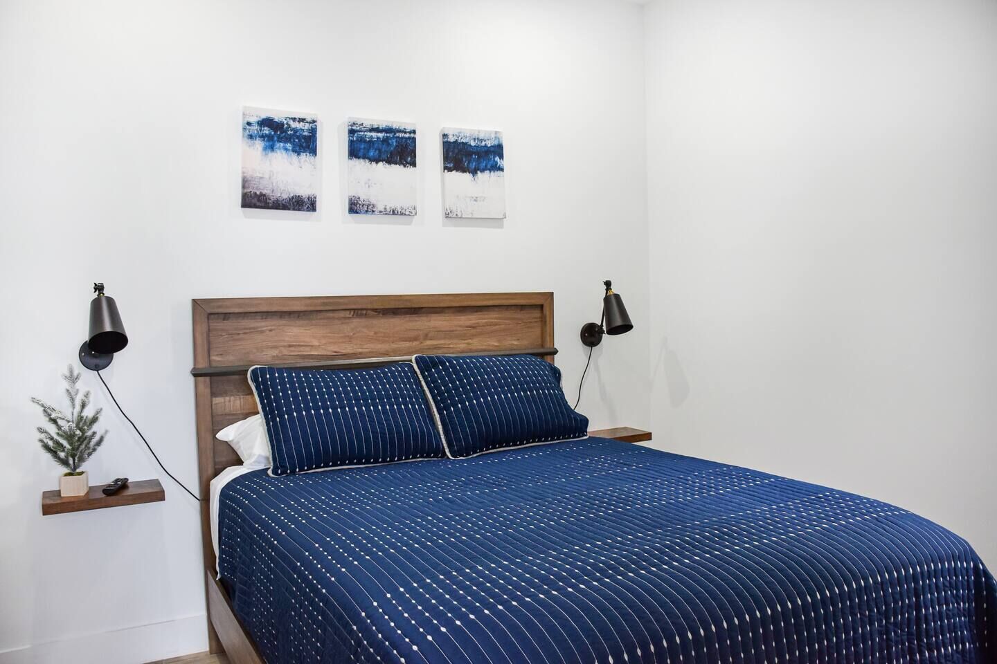 Une chambre à coucher avec une tête de lit en bois, une couvre-lit bleue à motifs, deux lampes de chevet et trois tableaux accrochés au mur.