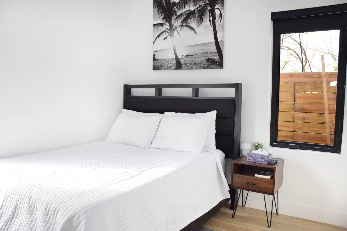 Uma cama com roupa de cama branca, um cabeceiro escuro, uma mesa de cabeceira com um controle remoto e uma janela com persiana de madeira.