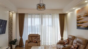 Ein modernes Wohnzimmer mit einer Couch, einem Sessel und einem Couchtisch. An der Wand ist ein Fernseher angebracht, es gibt eine Pflanze und ein Gemälde.
