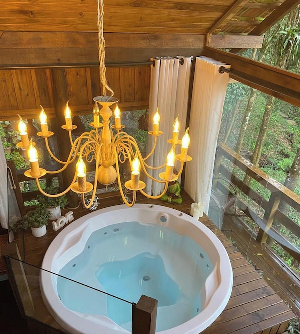 A wooden deck with a hot tub, a chandelier, and a view of greenery.