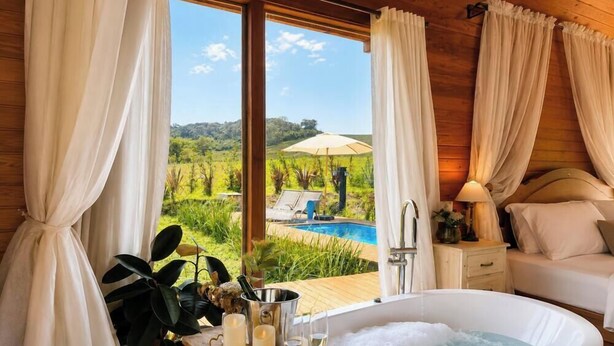 A bedroom with a view of a swimming pool, a bed, a bedside table with a lamp, and a window with curtains. - Cabanas da Mata - Alfredo Wagner - SC (Arnópolis)