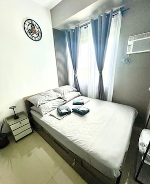 A bedroom with a bed, curtains, a wall clock, a bedside table, and an air conditioning unit. - Your Perfect Dasmarinas Getaway (Dasmarinas)