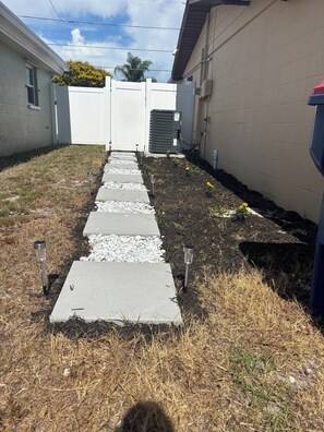 A pathway leading to a house with a white fence, an air conditioning unit, and a small garden area with gravel and grass. - Home Away From Home – Holiday, FL (Holiday)