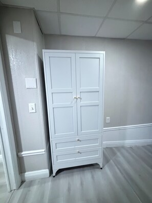 A white wardrobe with two doors and a lower shelf, positioned against a wall with a light gray finish and a wooden floor. - Home Away From Home – Holiday, FL (Holiday)