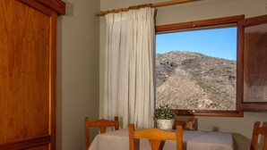 Un comedor con una mesa dispuesta para dos personas, sillas de madera, una ventana con vistas a un paisaje montañoso y una cortina.