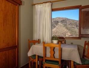 Ein Essbereich mit Tisch für zwei Personen, Holzsesseln, einem Fenster mit Blick auf eine Berglandschaft und einem Vorhang.