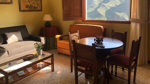 Un cuarto acogedor con un sofá, una mesa de comedor con sillas, una lámpara, una ventana con vistas a un paisaje montañoso y dos obras de arte enmarcadas en la pared.
