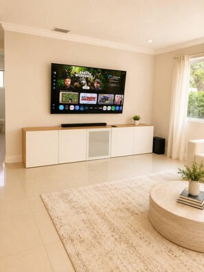 Ein modernes Wohnzimmer mit einem Wand-Fernseher, einem weißen Schrank darunter, einem runden Couchtisch mit Pflanze und einem großen Fenster mit Vorhängen.