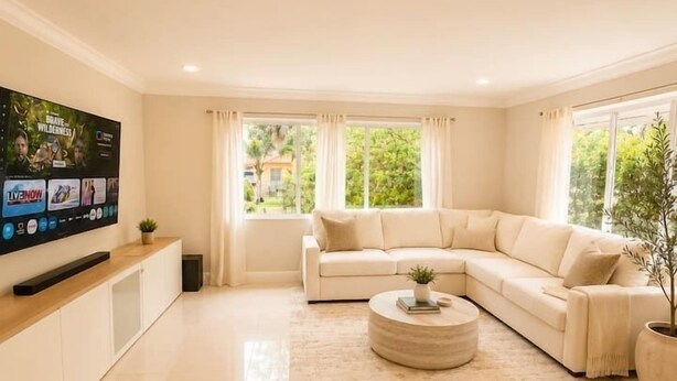 A modern living room with a beige sofa, a round coffee table, a flat-screen TV, and a large window with curtains. - Private Pool Oasis Near Wynwood, Midtown & Beaches (Miami)