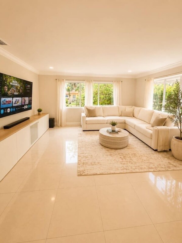 Ein modernes Wohnzimmer mit einem beigen Sofa, einem runden Couchtisch, einem Flachbildfernseher und einem großen Fenster mit Vorhängen.
