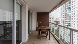 Un balcón con mesa y sillas de madera, un ventanal de vidrio y vistas a edificios altos.