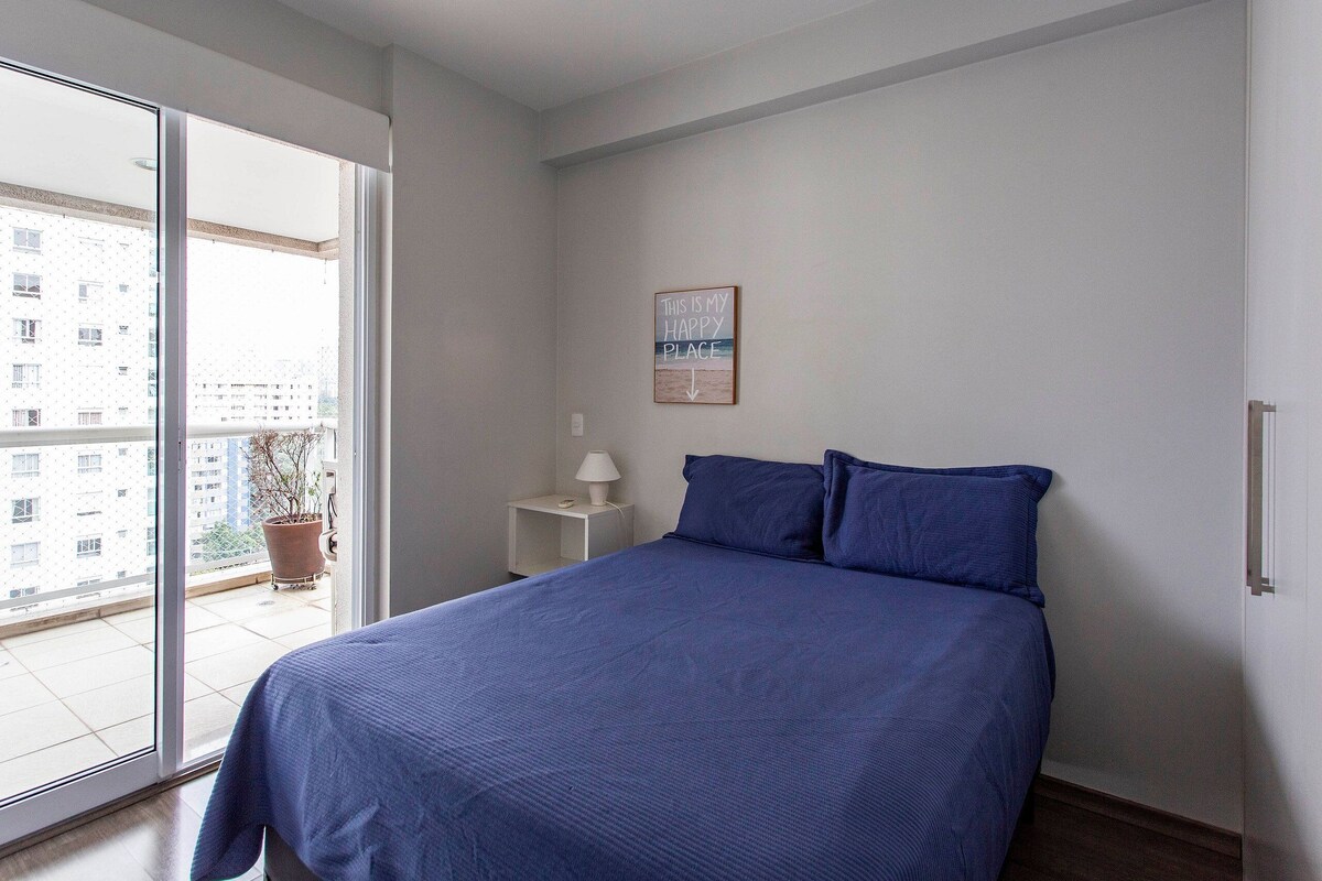 Une chambre à coucher avec un lit, une table de chevet, une lampe, une plante en pot et une vue sur un immeuble par la fenêtre.