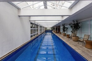 Une longue piscine intérieure aux parois en verre transparent et dotée d’une verrière au-dessus, entourée de plantes en pot et de sièges.