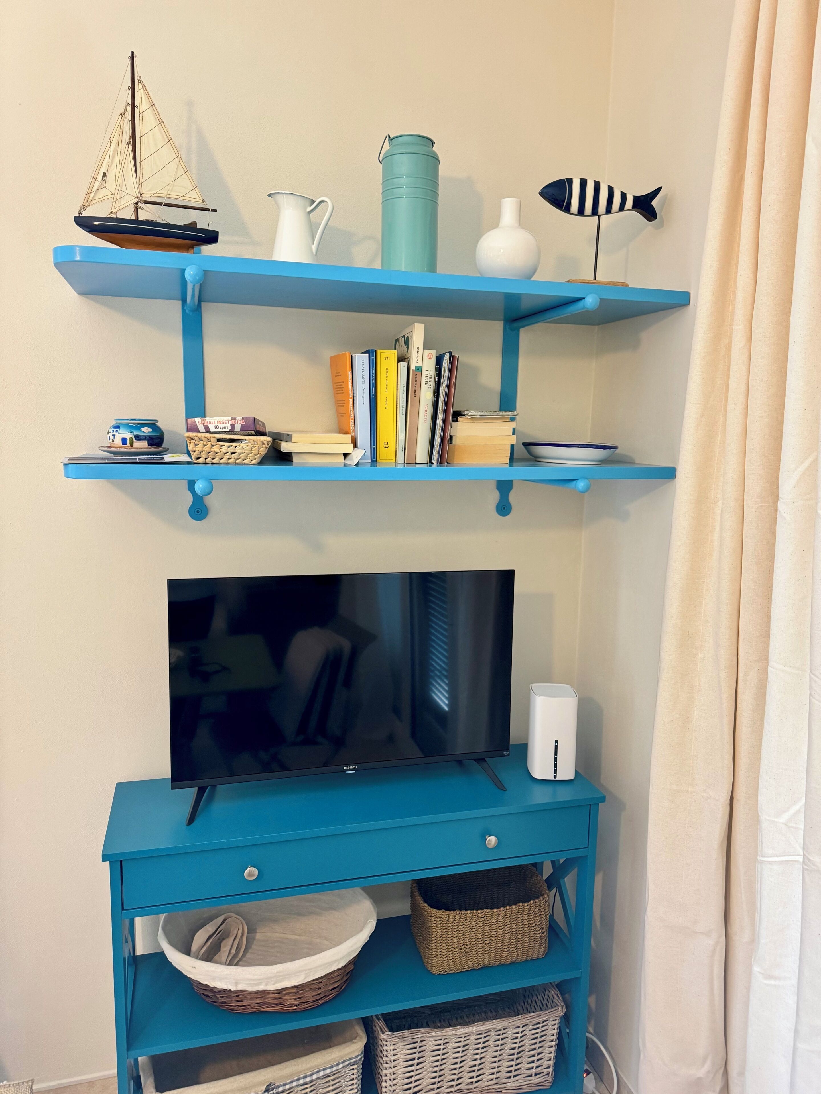 A room with a blue TV stand, a flat-screen TV, and a shelf with books, a teapot, and a small sailboat model.