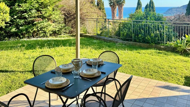An outdoor patio with a table set for dining, surrounded by chairs, and a view of the sea and hills in the background. - The House with a Blue Garden and Ocean View (Monade Santa Lucia)