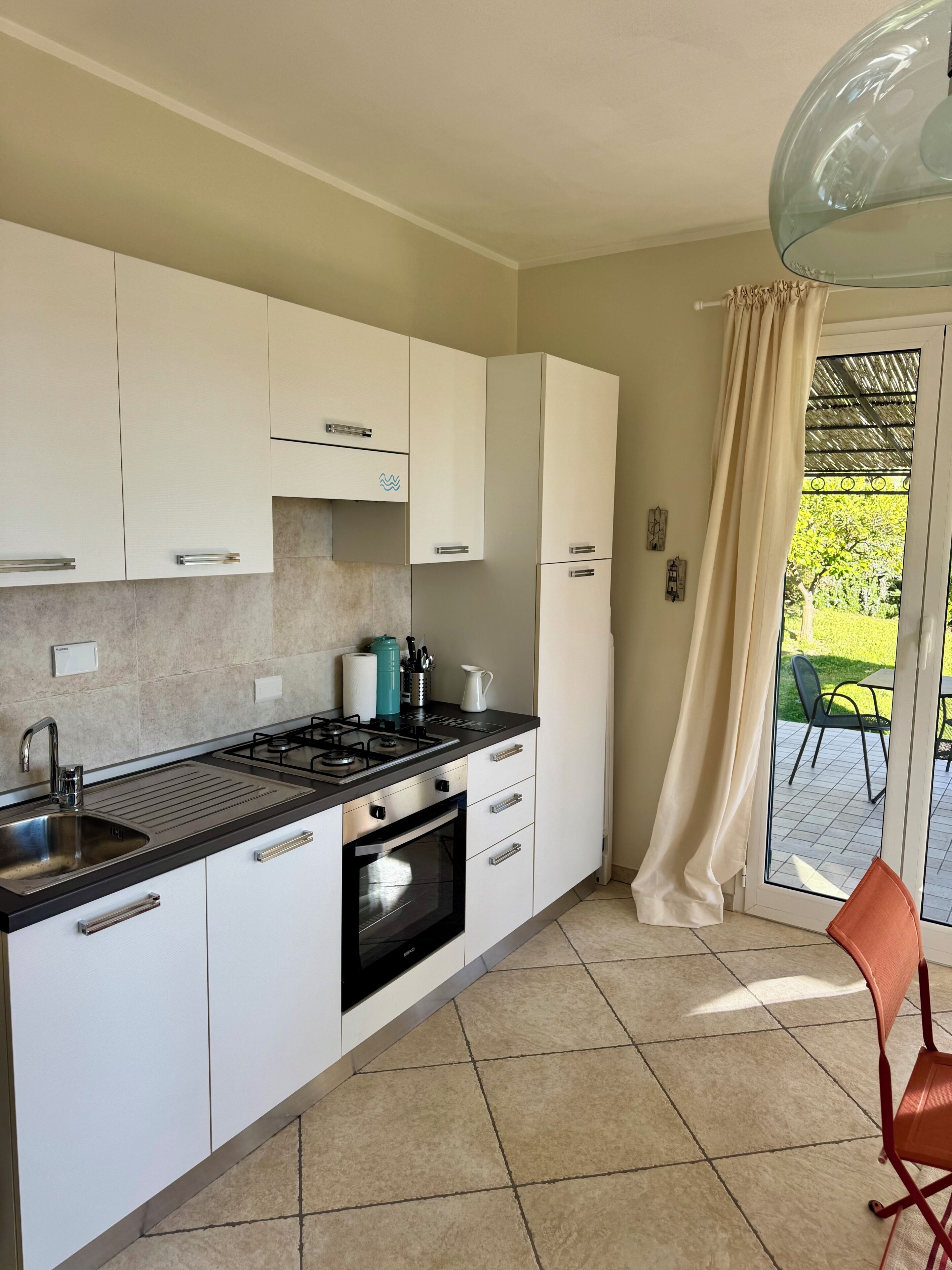 Una cucina moderna con mobili bianchi, piano cottura a gas, lavello e porta vetrata che dà su un patio esterno.