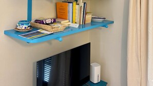 A room with a TV, shelves with books and decorative items, a small table with a basket, and a curtain. - The House with a Blue Garden and Ocean View (Monade Santa Lucia)