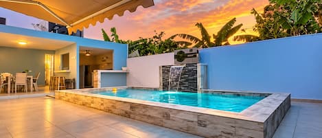 Ein Schwimmbecken mit Wasserfall, umgeben von einer modernen Terrasse mit überdachter Lounge-Ecke.