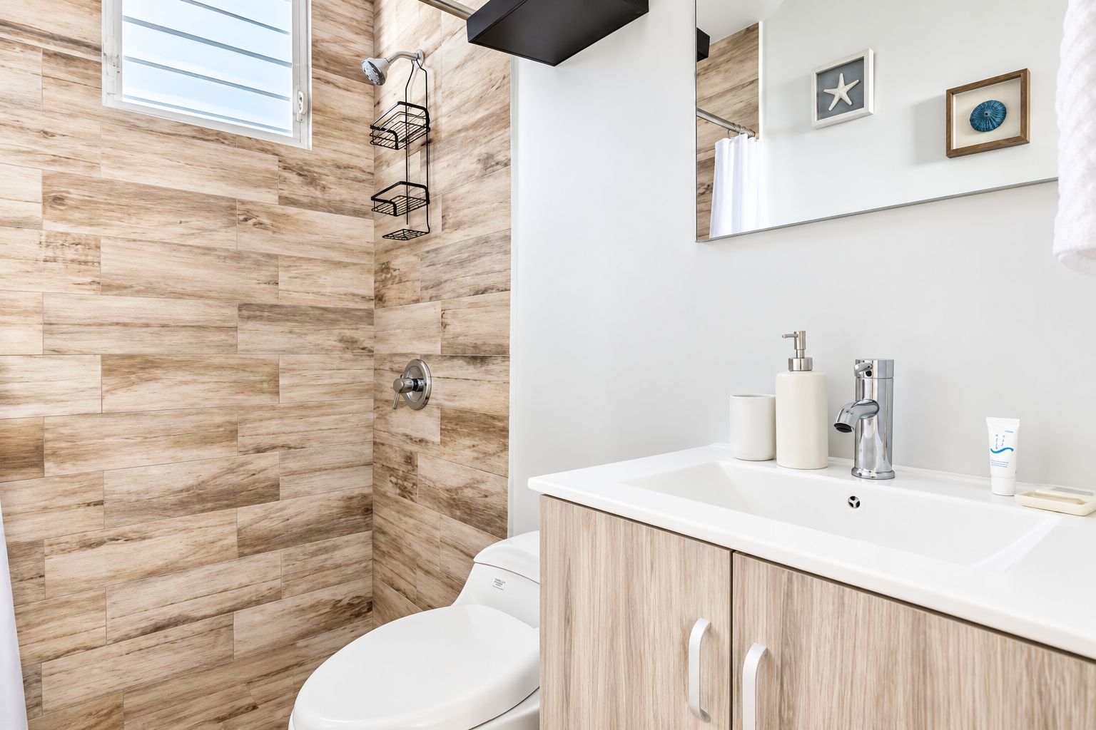 Un baño moderno con suelo de madera en espiga, ducha con mampara de vidrio, inodoro blanco y tocador con espejo y artículos de aseo.