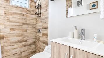 Un baño moderno con suelo de madera en espiga, ducha con mampara de vidrio, inodoro blanco y tocador con espejo y artículos de aseo.